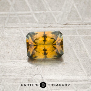 A rectangular, faceted orange sapphire weighing 0.70 carats rests on a textured surface with the “EARTH’S TREASURY” text and logo below it.