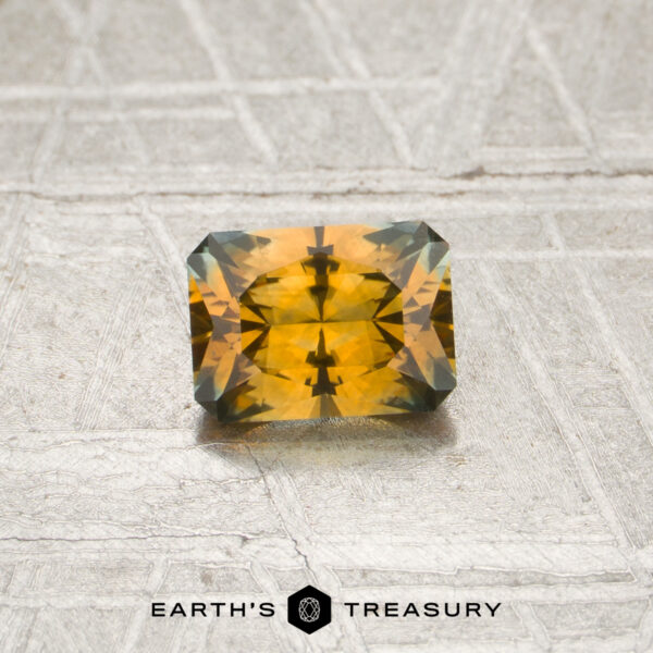 0.70-Carat Orange Montana Sapphire (Heated) A rectangular, faceted orange sapphire weighing 0.70 carats rests on a textured surface with the “EARTH’S TREASURY” text and logo below it.