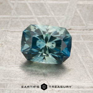 A 1.74-carat aqua-blue particolored Montana sapphire (heated), cut in a rectangular faceted shape, is displayed on a textured gray surface with the “Earth’s Treasury” logo below.