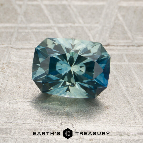 1.74-Carat Aqua-Blue Particolored Montana Sapphire (Heated) A 1.74-carat aqua-blue particolored Montana sapphire (heated), cut in a rectangular faceted shape, is displayed on a textured gray surface with the “Earth’s Treasury” logo below.