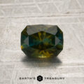 A 1.52-carat olive-yellow-blue particolored Montana sapphire (heat treated) rests on a textured, metallic surface above the “Earth’s Treasury” logo.