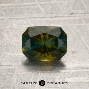 A 1.52-carat olive-yellow-blue particolored Montana sapphire (heat treated) rests on a textured, metallic surface above the “Earth’s Treasury” logo.