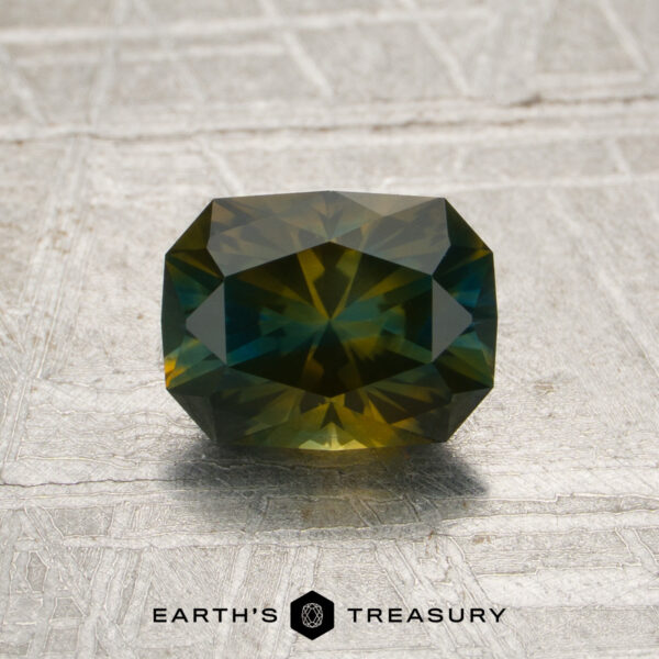 1.52-Carat Olive Green-Yellow-Blue Particolored Montana Sapphire (Heated) A 1.52-carat olive-yellow-blue particolored Montana sapphire (heat treated) rests on a textured, metallic surface above the “Earth’s Treasury” logo.