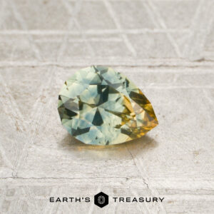A faceted pear-shaped 0.82-carat aqua-yellow particolored Montana sapphire (heated) with green and yellow hues rests on a textured surface above the text “Earth’s Treasury.”.