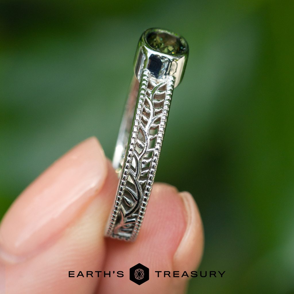 A hand holds the _DSC0652-1200-BW silver ring, featuring intricate engraving on the band and a green gemstone, set against a blurred green background.