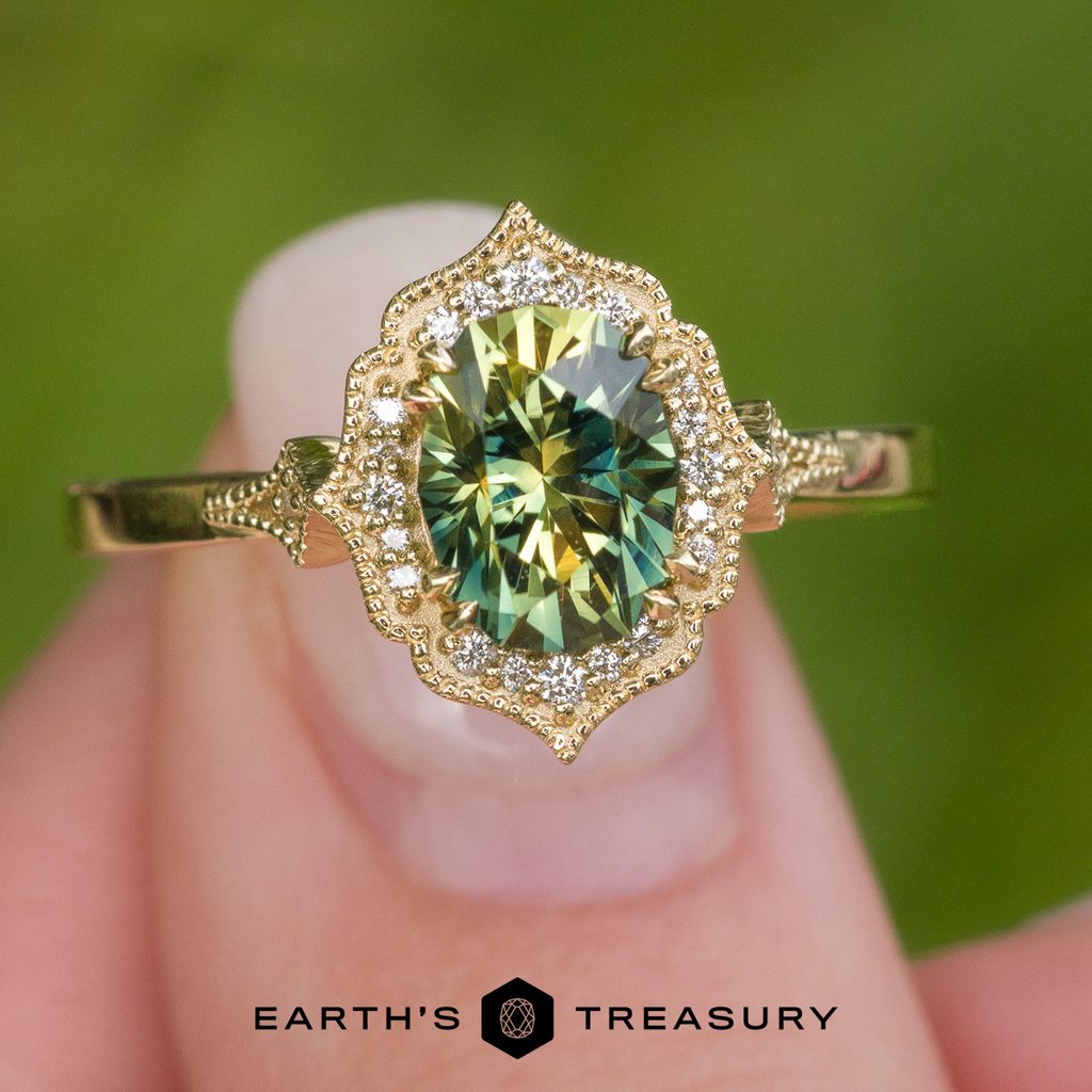 A person holds _DSC1957-1200-BW, a gold textured ring featuring an oval green gemstone surrounded by a halo of small diamonds, between two fingers. The “Earth’s Treasury” logo is displayed at the bottom.