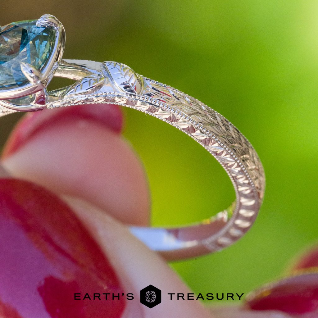 A close-up of the _DSC4528-1200-BW ring showcases its silver texture, intricate filigree engravings, and a blue gemstone, held between fingers with red nail polish. The Earth’s Treasury logo appears at the bottom.