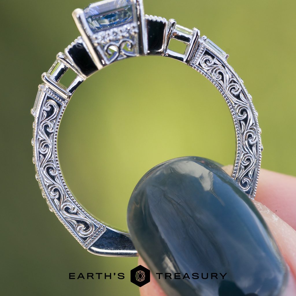 A close-up of the _DSC4908-1200-BW ring showcases its ornate silver design, featuring intricate filigree patterns and rich texture, held between painted fingertips against a softly blurred green background. The Earth’s Treasury logo appears at the bottom.