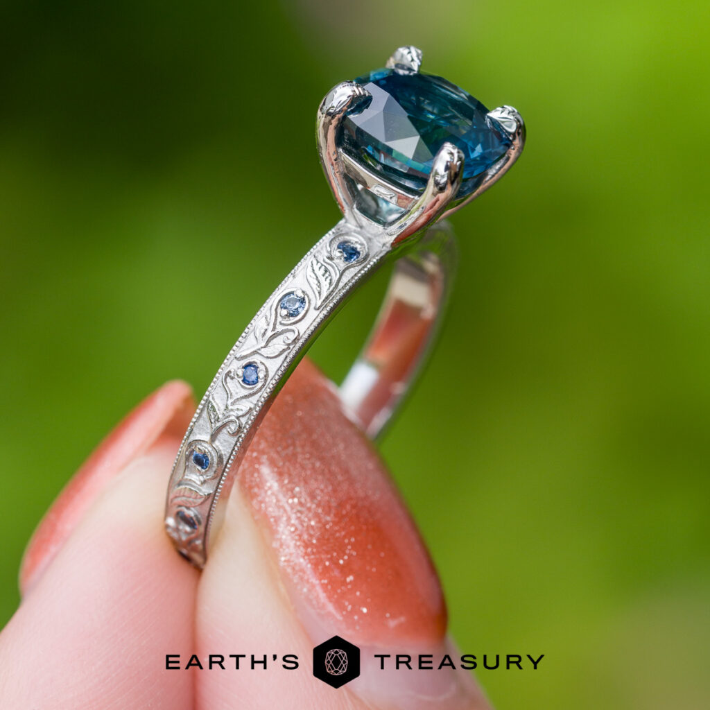 A hand displays a custom ring, featuring engraved nature-inspired patterns, a blue gemstone center, and small blue stones set along the band, against a softly blurred green background.