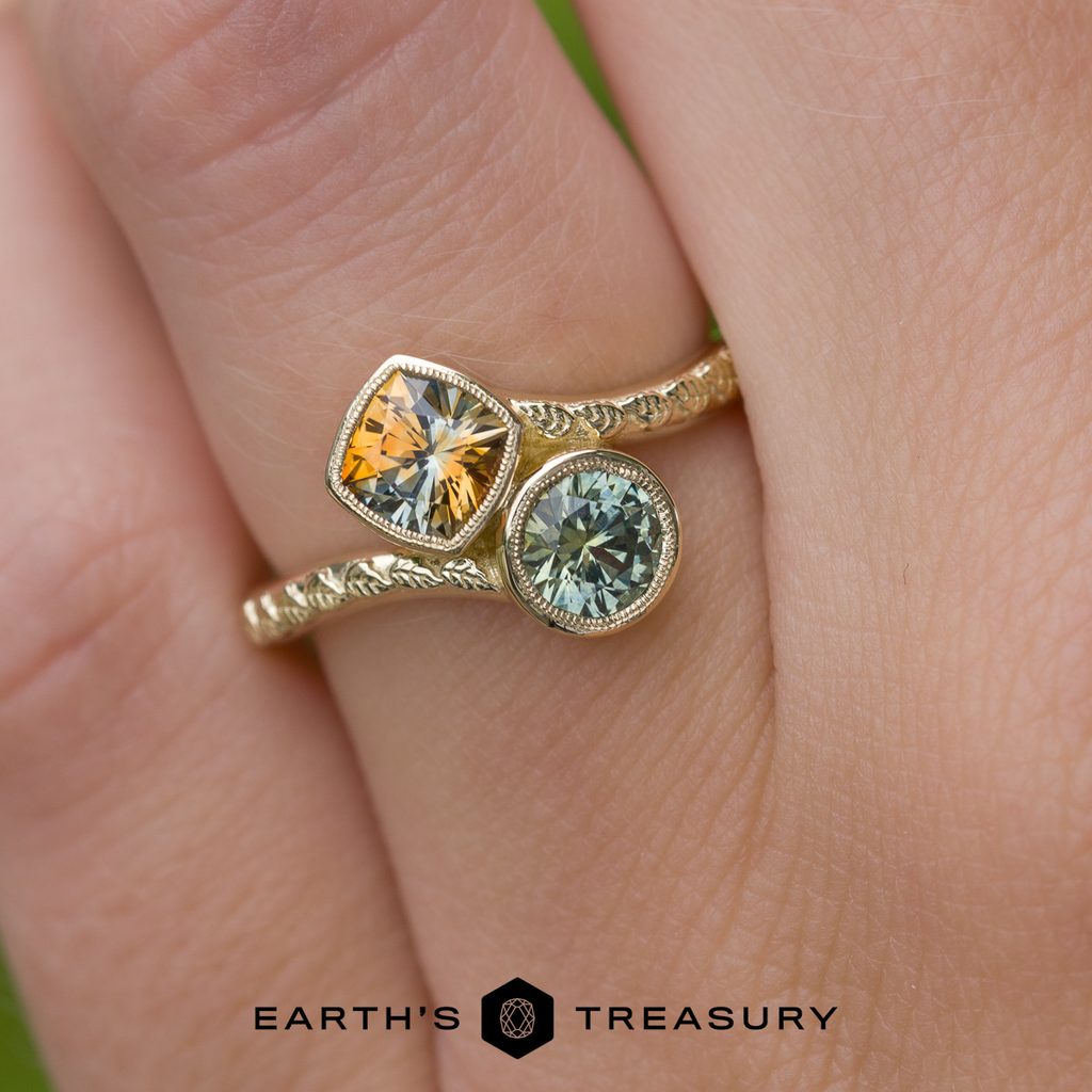 A hand wears the _DSC6103-1200-BW ring, a gold band adorned with one yellow and one green gemstone and delicate filigree detailing along the band. The “Earth’s Treasury” logo and text are displayed at the bottom of the image.