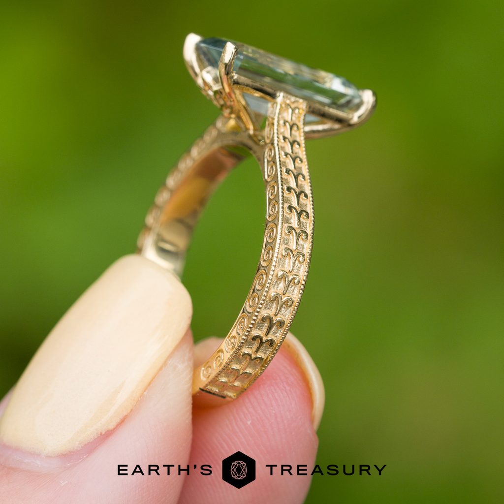 A hand holding the _DSC6433-1200-BW ring, featuring intricate filigree patterns and a large rectangular gemstone that highlights the exquisite texture of the piece, set against a soft green blurred background.