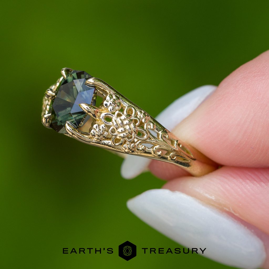 A hand with light blue nails displays the _DSC7069-1200-BW ring—an ornate gold band set with a dark green gemstone, featuring intricate filigree and delicate floral texturing. The “EARTH’S TREASURY” logo appears at the bottom.