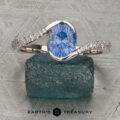 The "Cara" ring—featuring an oval blue gemstone accented by small diamonds set in silver—rests on a textured blue stone, showcased atop a patterned metal surface.