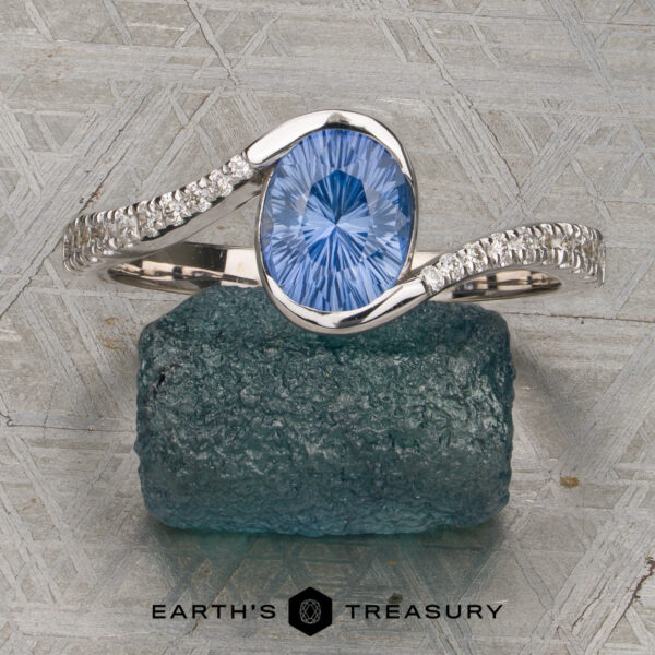 The "Cara" ring The "Cara" ring—featuring an oval blue gemstone accented by small diamonds set in silver—rests on a textured blue stone, showcased atop a patterned metal surface.