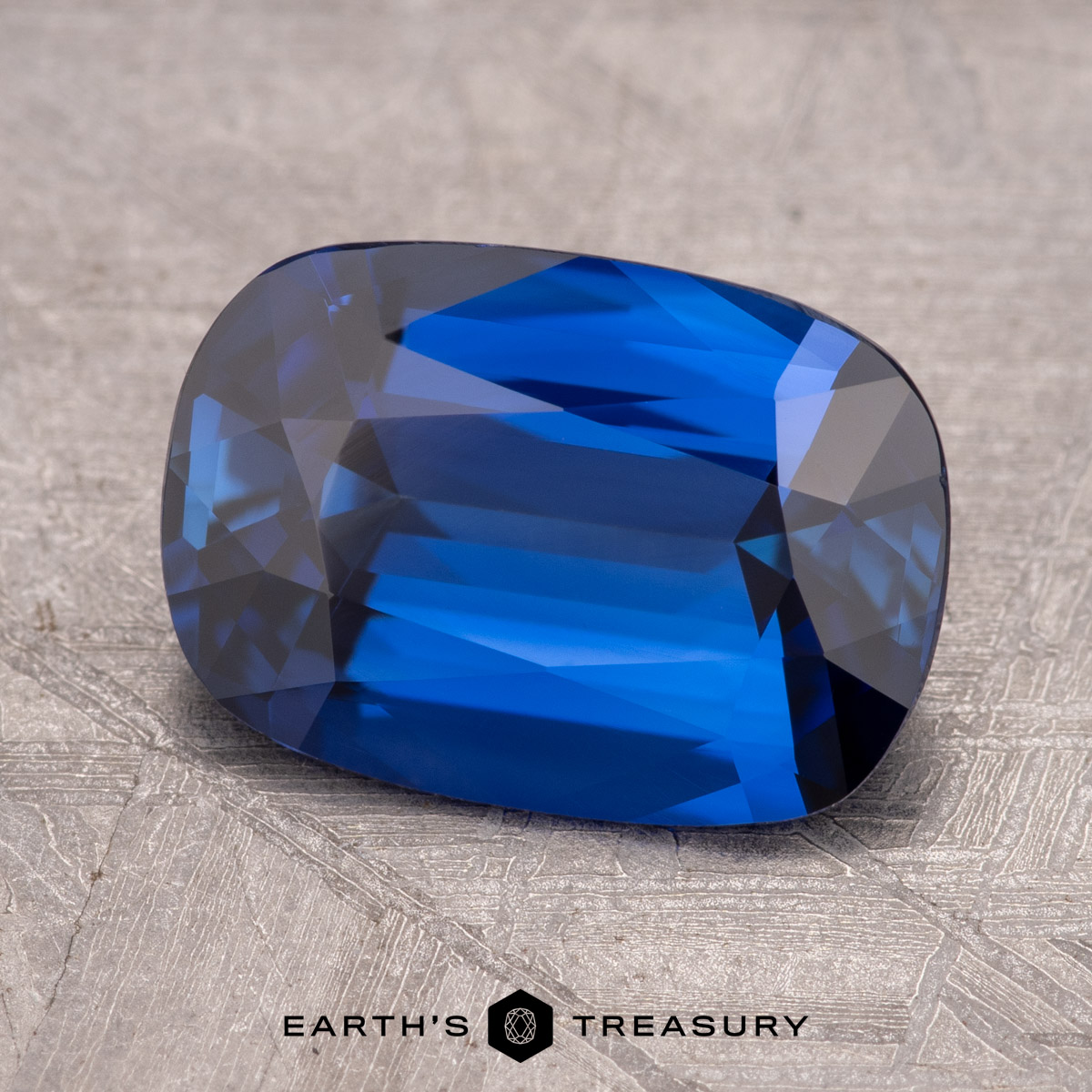 A deep blue, cushion-cut gemstone, SNY1767-1200, rests on a textured gray surface with the words “Earth’s Treasury” beneath it.