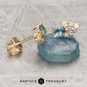 The Sapphire Pear Cluster Earrings with blue and white gemstones is displayed next to a matching gemstone and a gold earring back on a patterned gray surface, with the Earth’s Treasury logo at the bottom.