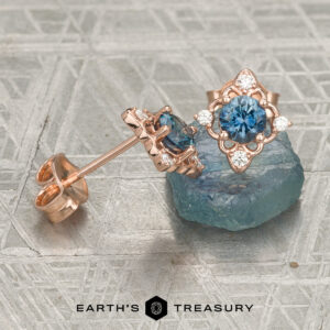 The "Marisol" Stud Earrings in 14K Rose Gold featuring blue gemstones and small white diamonds displayed on a blue crystal, with the text “Earth’s Treasury” at the bottom.