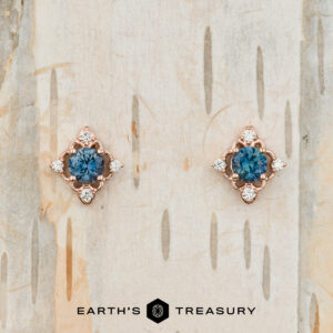 The "Marisol" Stud Earrings featuring blue gemstones and small diamonds set in rose gold, displayed on a light-colored textured surface.