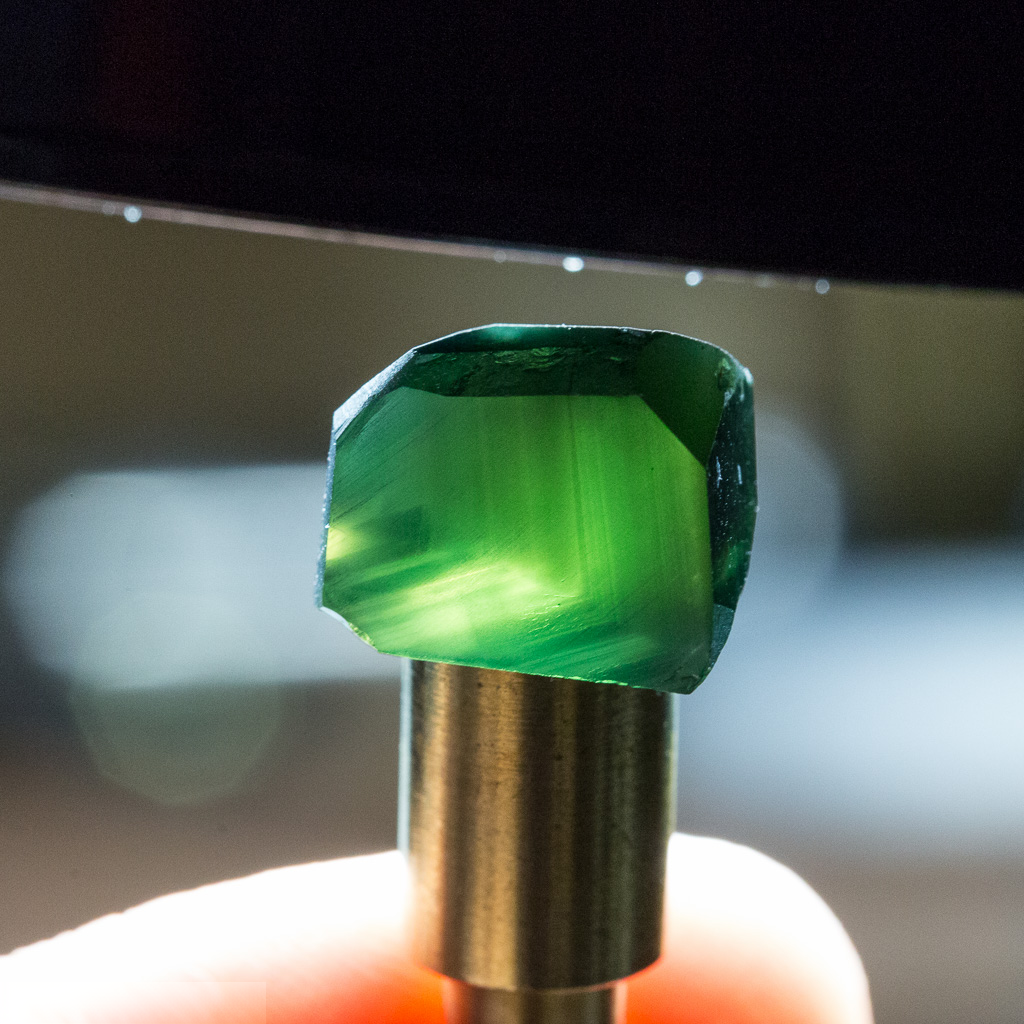 A green faceted gemstone is held upright in a metal holder, illuminated by bright light from behind—a captivating example of the brilliance showcased in “ethical-sourcing-what-does-it-mean.”.