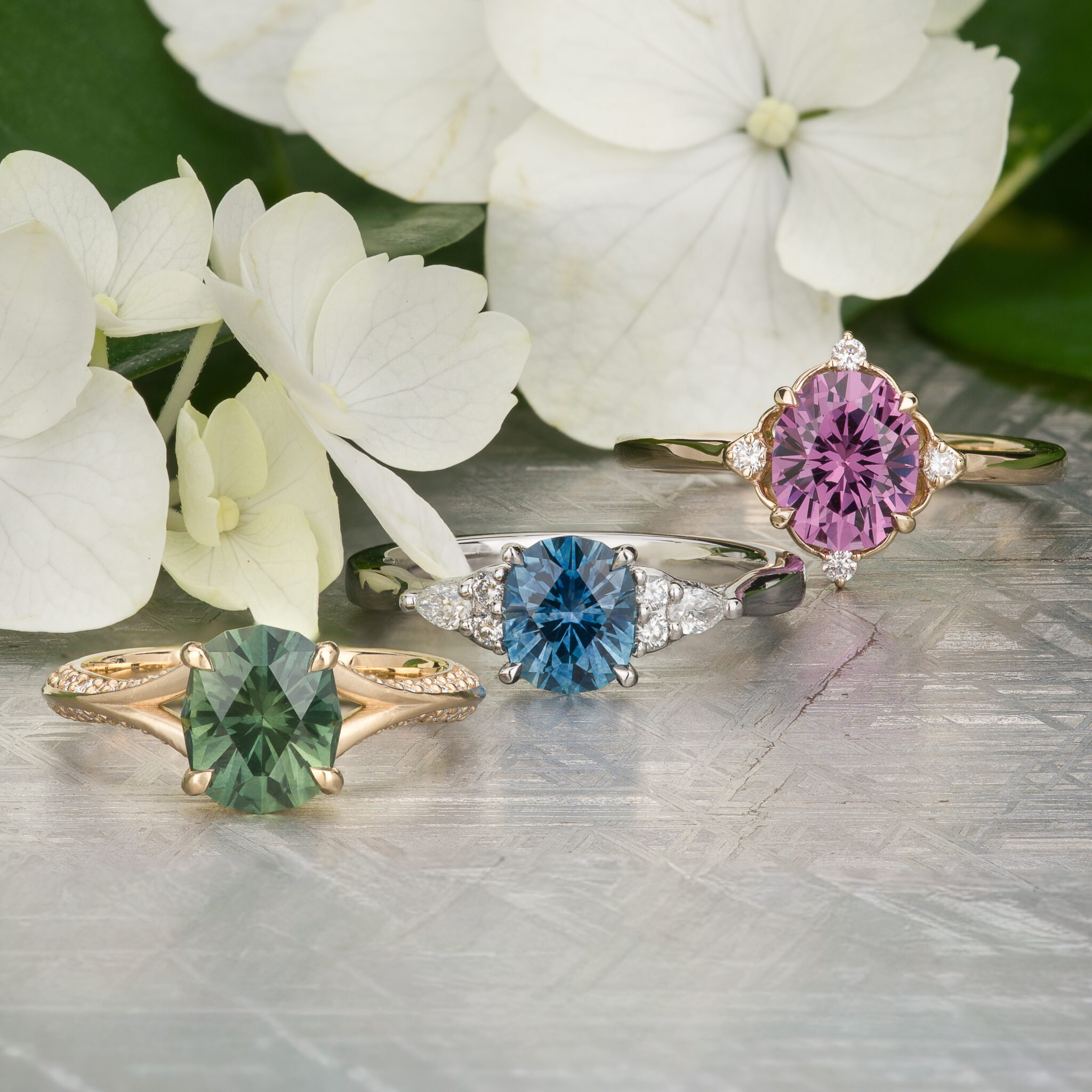 Three gemstone rings with green, blue, and purple stones—featuring stunning Australian Sapphires—are displayed on a patterned surface beside white hydrangea flowers. The image is titled “Stacked from 51 images. Method=B (R=14,S=1).”.