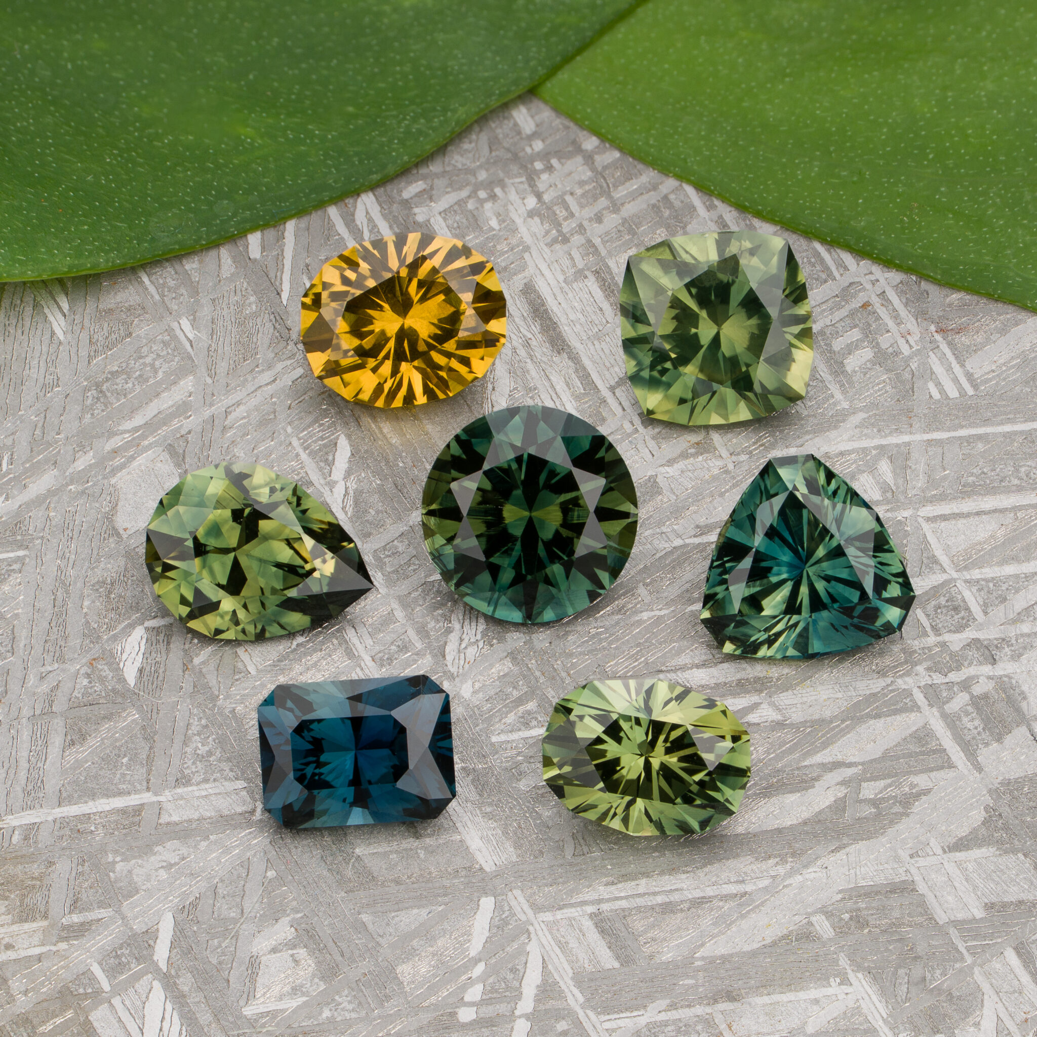 Here’s a rewritten version of the sentence incorporating your product data: Six faceted gemstones in shades of green, yellow, and blue—exceptional Australian Sapphires—are displayed on a textured surface with green leaves in the background. The image, titled “Stacked from 32 images. Method=B (R=7,S=2)”, captures their brilliance in stunning detail.